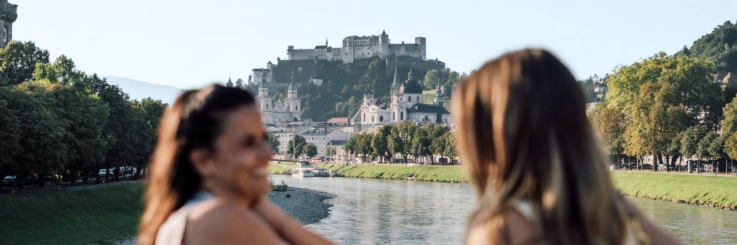 Due amici sul ponte Müllnersteg a Salisburgo, con la Fortezza di Hohensalzburg sullo sfondo. | © SalzburgLand Tourismus, Chris Perkles