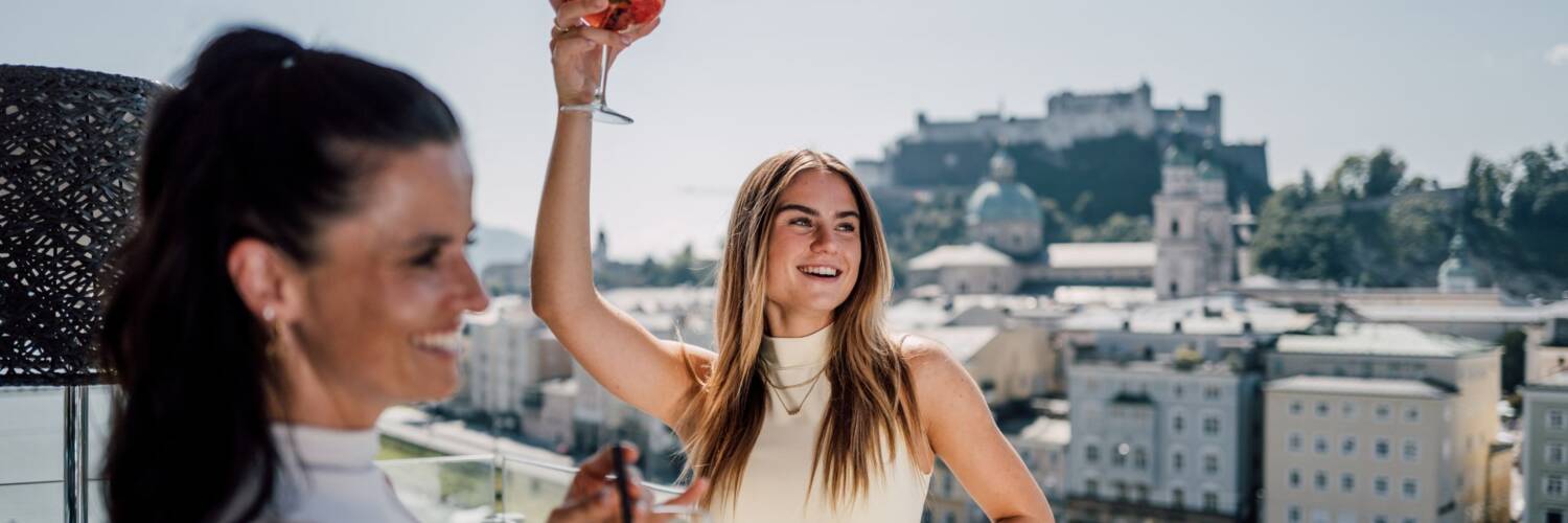Zwei Frauen mit Getränken mit der Festung Hohensalzburg im Hintergrund | © SalzburgerLand Tourismus, Chris Perkles