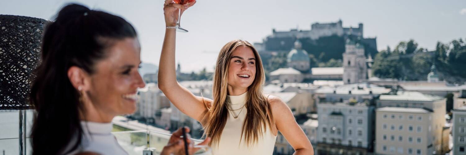 Two women with drinks and the Festung Hohensalzburg in the background | © SalzburgerLand Tourismus, Chris Perkles