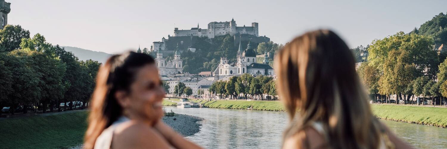 Two friends on Müllnersteg in Salzburg, with Hohensalzburg Fortress in the background. | © SalzburgLand Tourismus, Chris Perkles