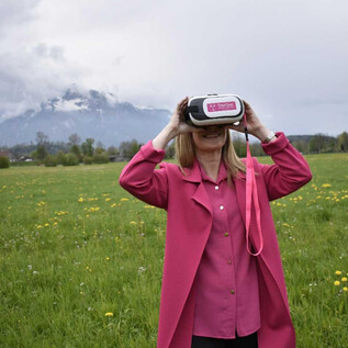 A woman is wearing a virtual reality headset and looking into the distance. In the background, there are green meadows and mountains. | © Sabine Rath