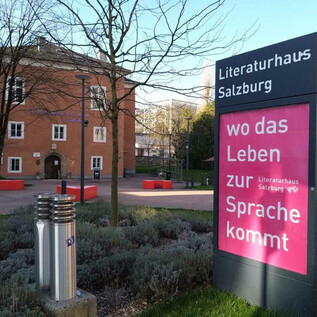 Dom Literatury w Salzburgu to nowoczesny budynek z przyjaznym otoczeniem na zewnątrz. Obok głównego biura znajduje się tablica informacyjna z napisem "gdzie życie staje się mową". | © Literaturhaus Salzburg