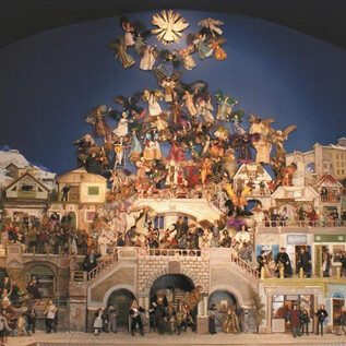 Una grande, dettagliata natività con molte figure e case sotto un cielo blu. La scena è riccamente decorata e mostra un'atmosfera festosa. | © Textil-Kunst-Galerie Sammlung Aichhorn