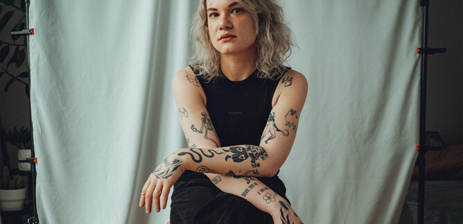 A person with a tattoo covering sits on a chair against a white background. They have curly blonde hair and wear a black top and pants. | © Yannic Steuerer