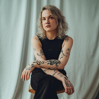 Eine Person mit einer Tattoo-Bedeckung sitzt auf einem Stuhl vor einem weißen Hintergrund. Sie hat lockige, blonde Haare und trägt ein schwarzes Oberteil und Hosen. | © Yannic Steuerer