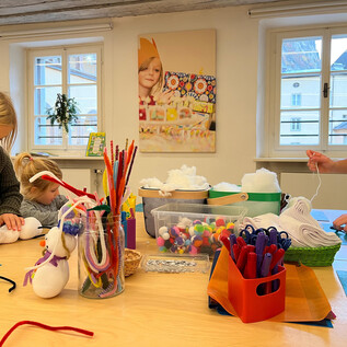 Zwei Kinder basteln mit bunten Materialien am Tisch, während eine Erwachsene ihnen hilft. Der Raum ist hell und freundlich, mit vielen Bastelutensilien verteilt. | © Altstadt Salzburg