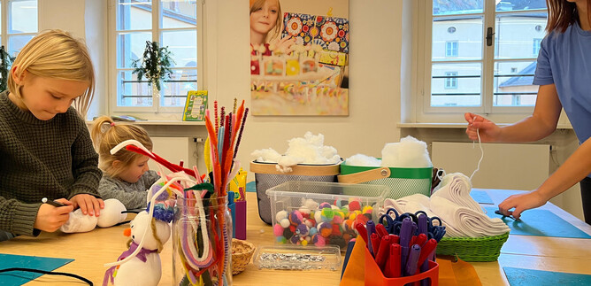 Two children are crafting with colorful materials at the table, while an adult helps them. The room is bright and cheerful, with many crafting supplies scattered about. | © Altstadt Salzburg
