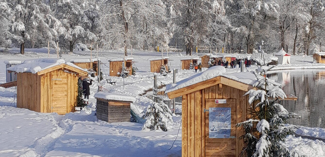 Zimowy krajobraz z małymi drewnianymi chatkami wokół zamarzniętego jeziora. Niebo jest czyste, a drzewa pokryte są śniegiem. | © Adventmarkt am Salzachsee