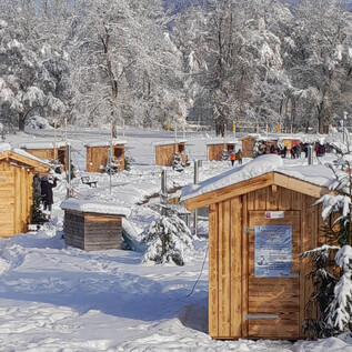 Eine verschneite Landschaft mit kleinen Holzhütten um einen gefrorenen See. Der Himmel ist klar und die Bäume sind mit Schnee bedeckt. | © Adventmarkt am Salzachsee