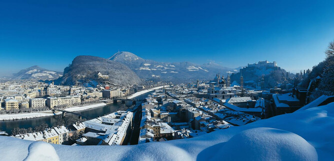 Ośnieżony krajobraz miejski z górami w tle i czystym niebieskim niebem. Dachy pokryte są śniegiem, a rzeka wiję się przez miasto. | © TSG_Breitgger