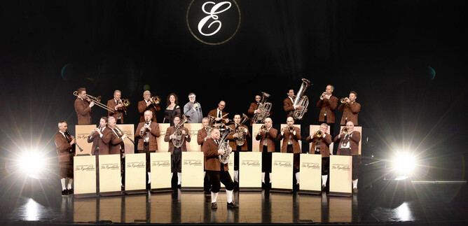 Une grande fanfare se tient sur la scène dans des costumes bruns élégants. Des projecteurs éclairent les musiciens pendant qu'ils jouent de leurs instruments. | © Paul Gärtner