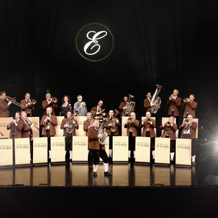 A large brass band stands on stage in elegant brown suits. Spotlights illuminate the musicians as they play their instruments. | © Paul Gärtner