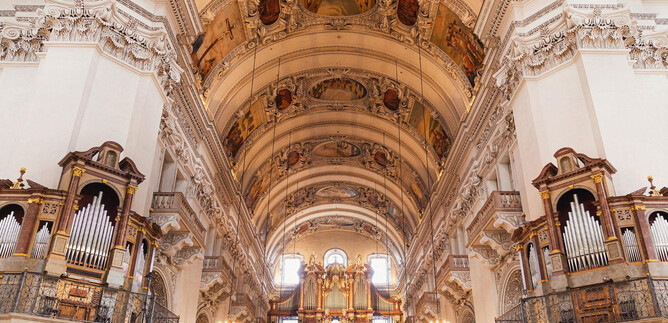 Eine beeindruckende Kircheninneneinrichtung mit hohen Gewölben und kunstvollen Deckengemälden. Auf beiden Seiten befinden sich große Orgeln und elegantes Licht beleuchtet den Raum. | © Erzdiözese Salzburg