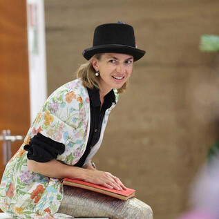 A woman with a tall hat sits smiling on a bench. She wears a colorful jacket and holds a book in her hand. | © Franz Neumayr