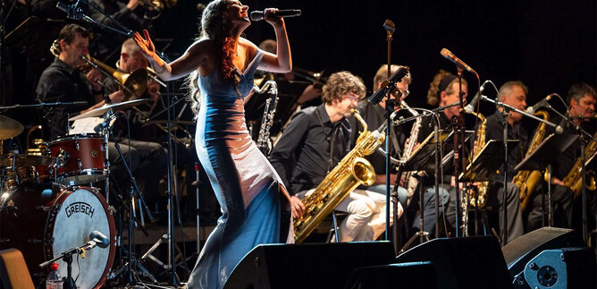 A singer in an elegant dress performs on stage in front of a band. In the background, musicians play various wind instruments. | © Albert Moser