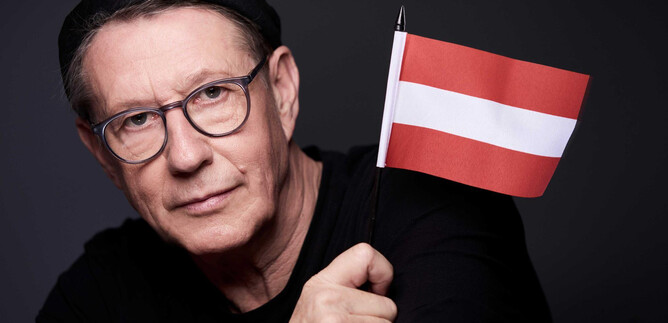 A man is holding a small red-and-white flag. He is wearing a black cap and glasses. | © Susie Knoll