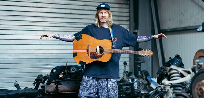 Un homme avec de longs cheveux se tient dans un atelier entre de vieilles pièces de voiture. Il tient une guitare et porte des vêtements décontractés. | © Michael McKee