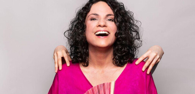 A woman with curly hair is wearing a bright pink top and holding a fan. In the background, two hands can be seen gently supporting her. | © Jasmin Schuller