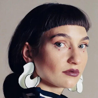 Eine Person mit kurzen, dunklen Haaren und auffälligen, weißen Ohrringen blickt direkt in die Kamera. Der Hintergrund ist neutral und hebt den Fokus auf das Gesicht. | © Anna Hadaier