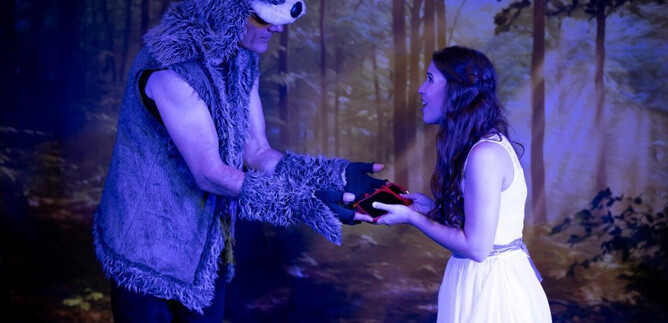 Ein Mann in einem Bärenkostüm übergibt einer Frau in einem weißen Kleid einen roten Gegenstand. Die Szene spielt in einem mystischen Wald. | © Theater mit Seele