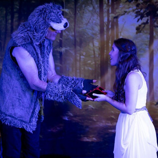 Un homme déguisé en ours remet à une femme en robe blanche un objet rouge. La scène se déroule dans une forêt mystique. | © Theater mit Seele