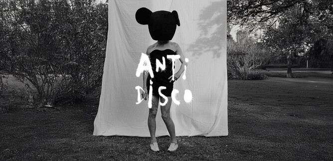 Eine Person mit einem großen, runden Kopf mit Ohren steht vor einem weißen Hintergrund. Der Schriftzug "ANTI DISCO" ist in der Mitte des Bildes zu sehen. | © Roman Goebel