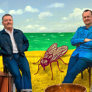 Zwei Männer sitzen in der Natur, umgeben von einer bunten Landschaft. Im Hintergrund sieht man das Meer und eine große Fliege aus Kunst. | © Helmut Wimmer