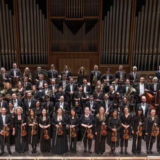 A large orchestra is standing on stage in formal attire. In the background, tall organ pipes can be seen. | © recreation Event GmbH
