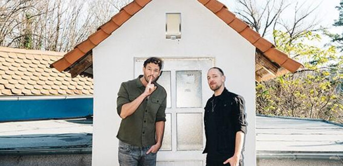 Two men are standing in front of a small, white house with a red roof. The surroundings are bright and there are trees in the background. | © Ingo Pertramer