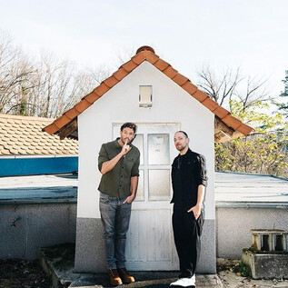 Two men are standing in front of a small, white house with a red roof. The surroundings are bright and there are trees in the background. | © Ingo Pertramer