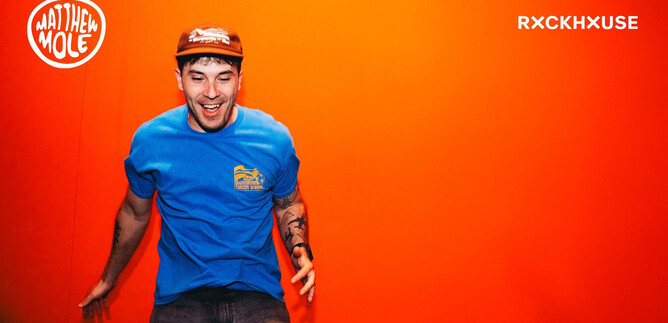 Mladý muž v modrém tričku a červené čepici stojí před oranžovou zdí a usmívá se. Vypadá veselý a energetický. | © Rockhouse Salzburg