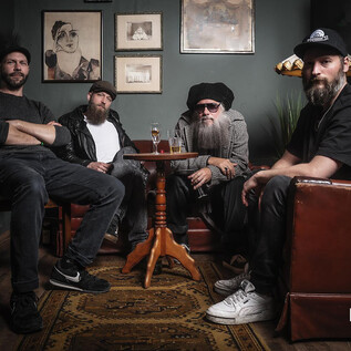 A group of four men is sitting in a cozy corner with a wooden chair and a table. The wall is painted dark and decorated with artwork. | © Photo Jockey
