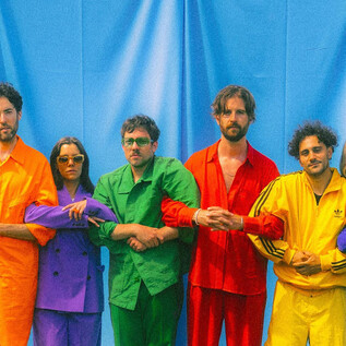 Hat személy áll egy kék háttér előtt, és színes ruhákat viselnek sárgában, zöldben, narancsban és lilában. Együtt pózolnak, és láthatóan vidám hangulatot árasztanak. | © Paul Vincent Schütz