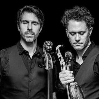 Two musicians stand close together, with serious expressions. They hold musical instruments in their hands and are depicted against a dark background. | © Stephan Doleschal