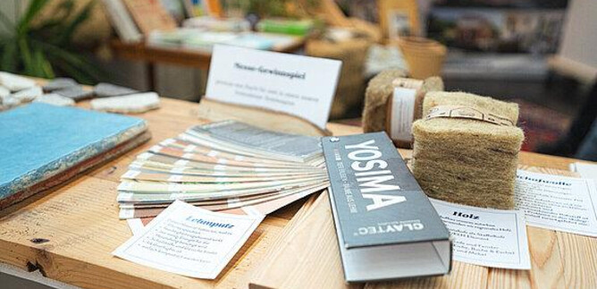 Ein Holztisch mit verschiedenen Materialien und Informationskarten. Darunter sind Farbmuster, Bücher und natürliche Materialien zur Verwendung in Bauprojekten. | © Messezentrum Salzburg