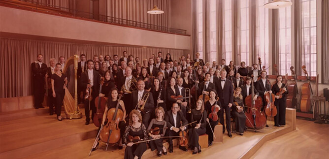 Ein großes Orchester steht in einem Raum mit hohen Fenstern. Die Musiker tragen formelle Kleidung und halten ihre Instrumente. | © Noë_Flum@