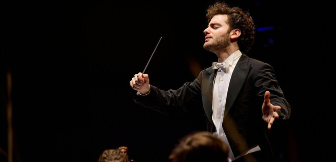 Ein dirigierender Musiker in einem eleganten Anzug leitet ein Orchester. Die Atmosphäre ist festlich und voller musikalischer Energie. | © Oliver Borchert