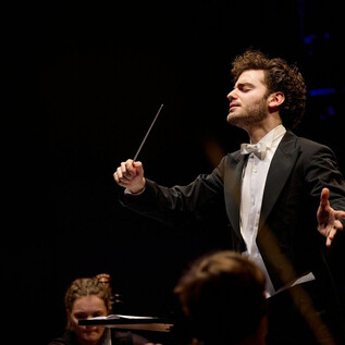 Ein dirigierender Musiker in einem eleganten Anzug leitet ein Orchester. Die Atmosphäre ist festlich und voller musikalischer Energie. | © Oliver Borchert