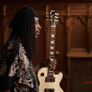 Egy zenész áll a kezében egy gitárral. Feltűnő ing van rajta, és gondolkodva néz a távolba. | © Tino Sieland