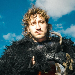 A man with curly hair is wearing a striking black fur coat. The background shows a clear sky with some clouds. | © Christopher Glanzl