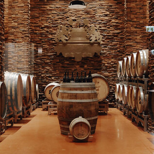 A cellar with wooden barrels and a stone wall. In the center stands a barrel with wine bottles. | © Sarah Matzer Photography / www.designmatzer.at
