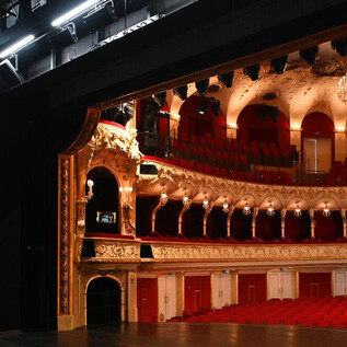 Ein Blick hinter die Bühne eines Theaters mit roten Sitzen und einer eleganten Einrichtung. Die Bühne ist beleuchtet und zeigt prächtige Details. | © Neumayr_Leo