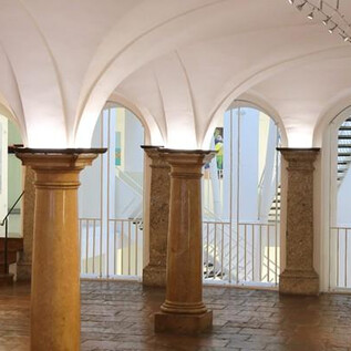 Una sala de exposiciones iluminada con columnas y obras de arte en las paredes. Al fondo se pueden ver escaleras y otros elementos de diseño. | © Stadtgalerie Rathaus