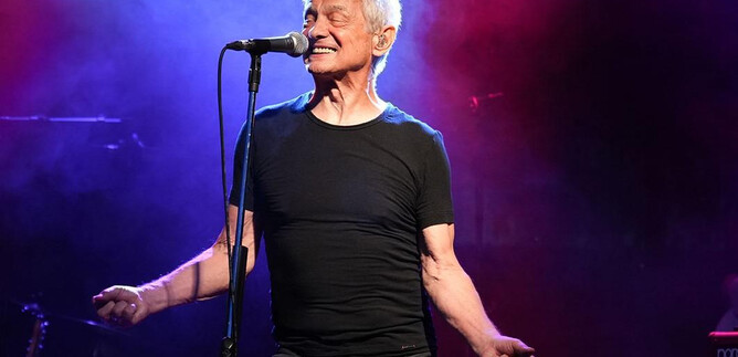 Ein älterer Mann steht auf der Bühne und singt in ein Mikrofon. Bunte Lichter und Nebel schaffen eine lebendige Konzertatmosphäre. | © Paul Trondl