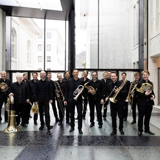 A group of musicians with brass instruments is standing in a modern room. They are wearing black clothing and smiling at the camera. | © Mozarteumorchester Salzburg
