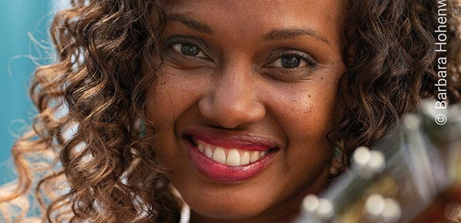 Egy mosolygó portré egy göndör hajú nőről, aki gitárt tart. A háttér elmosódott és egy kellemes hangulatot ábrázol. | © shutterstock / Barbara Hohenwallner