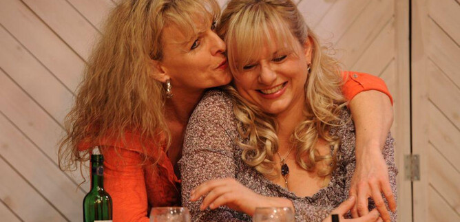 Zwei Frauen lachen und umarmen sich fröhlich an einem Tisch. Auf dem Tisch stehen Gläser, eine Flasche und Zigaretten. | © Heinz Bayer