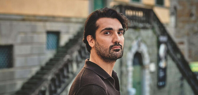 A man stands in front of a historic building with stairs in the background. He looks directly at the camera and has a thoughtful expression on his face. | © Ben Wolf