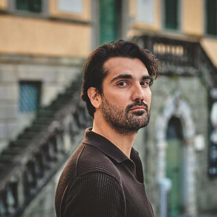 A man stands in front of a historic building with stairs in the background. He looks directly at the camera and has a thoughtful expression on his face. | © Ben Wolf