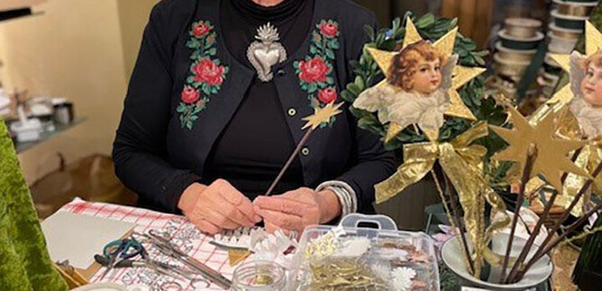 Eine freundliche ältere Frau sitzt an einem Tisch voller Bastelmaterialien. Um sie herum sind verschiedene Dekorationen und Materialien für das Handwerk zu sehen. | © Salzburger Heimatwerk